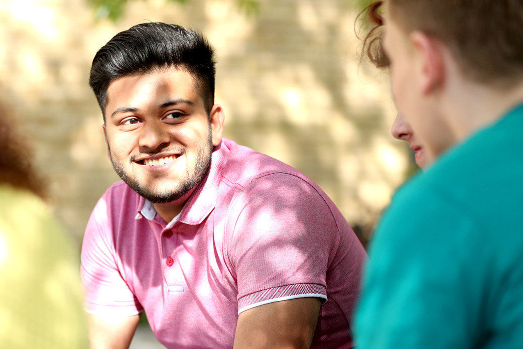 A smiling young man sits with friends under trees shaded from the sun outside the college